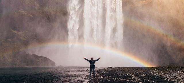 A person standing at the base of a waterfall under a rainbow.
